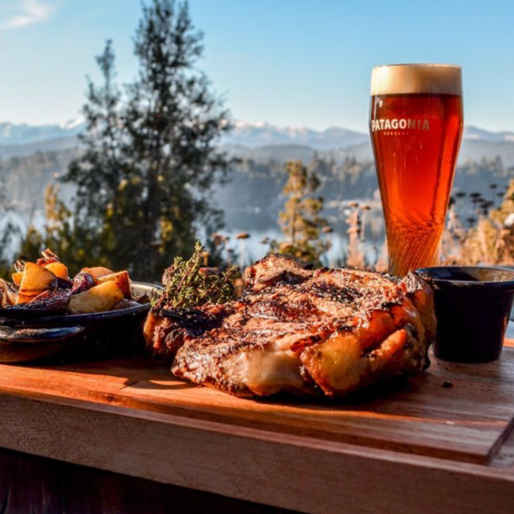 tábua com corte de carne e copo de cerveja com paisagem de lago e montanha de neve ao fundo